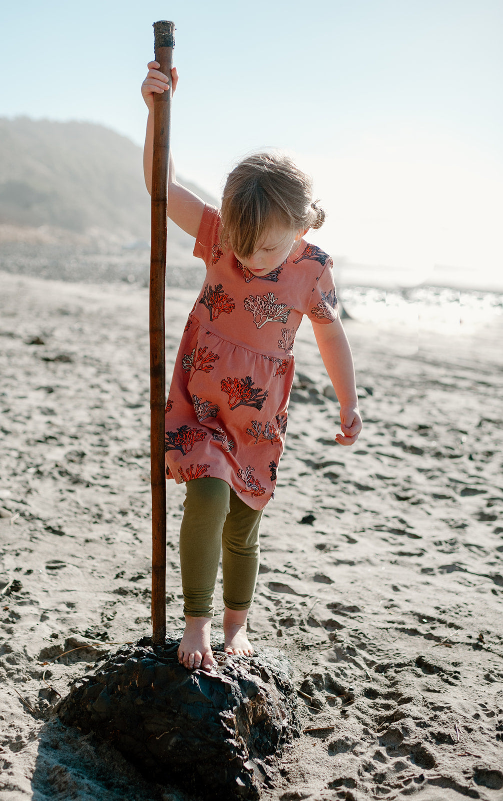 A young girl on the beach wearing this coral dress, standing with driftwood