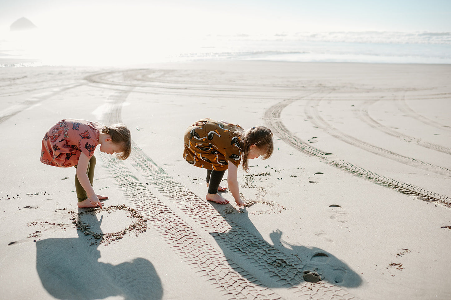 A photo of two young girls drawing in the sand on the beach, one in this coral dress